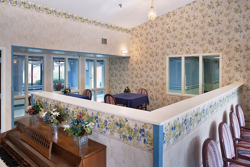 Interior common dining area with floral wallpaper, a piano, tables with purple tablecloths, and chairs around a half wall.
