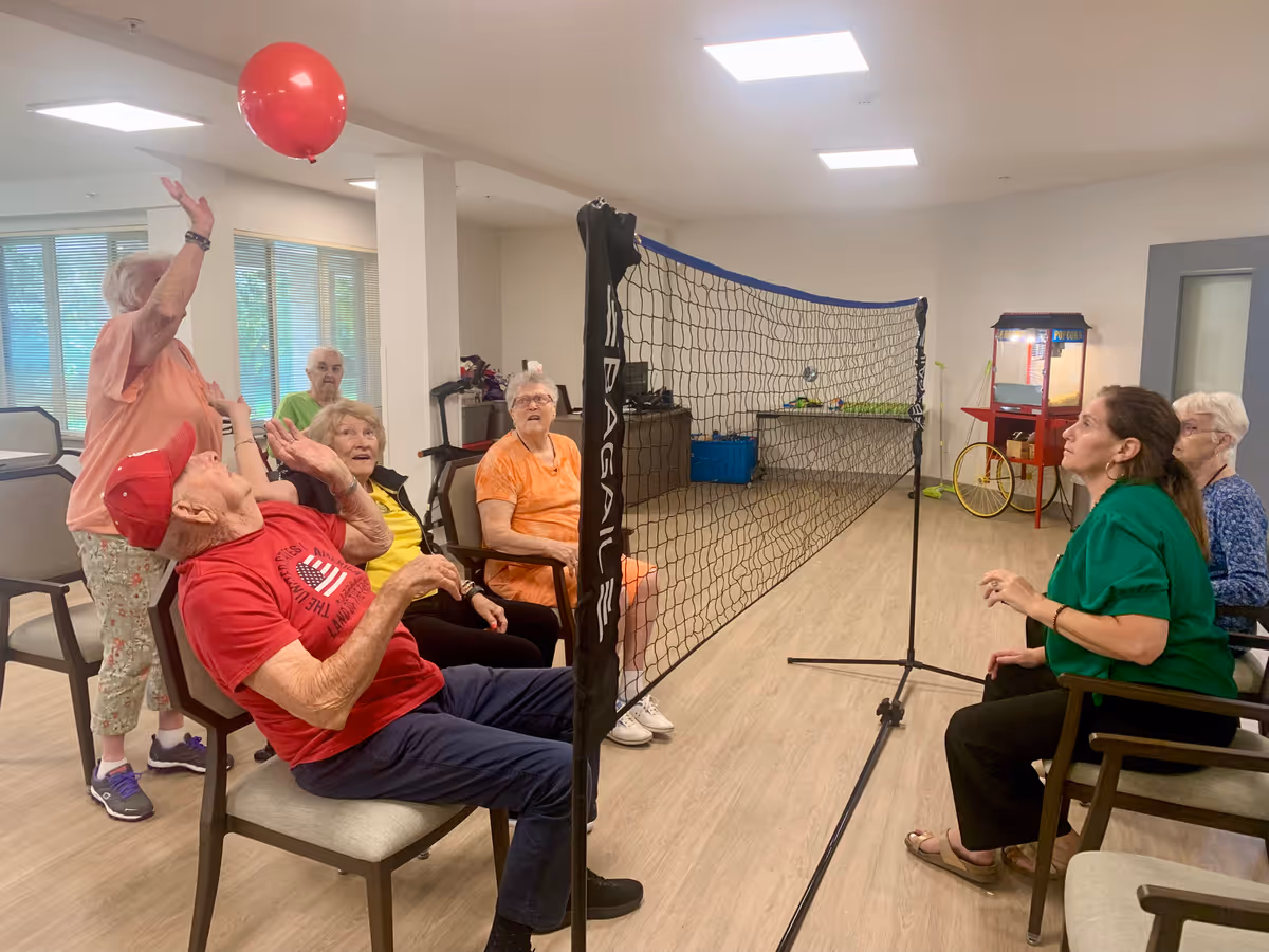 A group of elderly people seated in chairs playing a seated volleyball game indoors with a red balloon over the net. The room has large windows, light wood flooring, and a popcorn machine in the background.