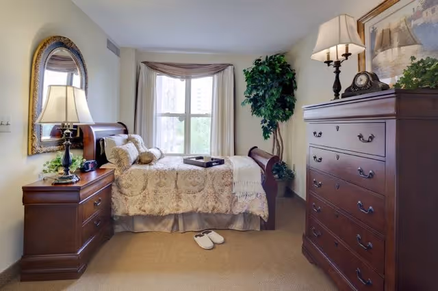 Cozy bedroom with a bed, nightstand and lamp, chest of drawers, window, and slippers on the floor.