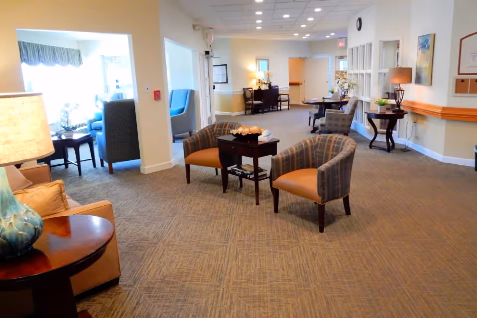 A spacious, well-lit common area in a senior living facility with several armchairs arranged around small tables, lamps providing warm lighting, and a carpeted floor. The area has a neutral color scheme with beige walls and white trim, and there are windows letting in natural light. The space appears comfortable and inviting for residents to sit and socialize.