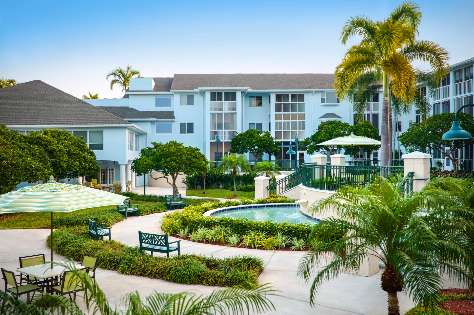 Outdoor courtyard area at Vi at Lakeside Village featuring a small pool surrounded by greenery, palm trees, benches, and tables with umbrellas, with a multi-story residential building in the background under a clear blue sky.