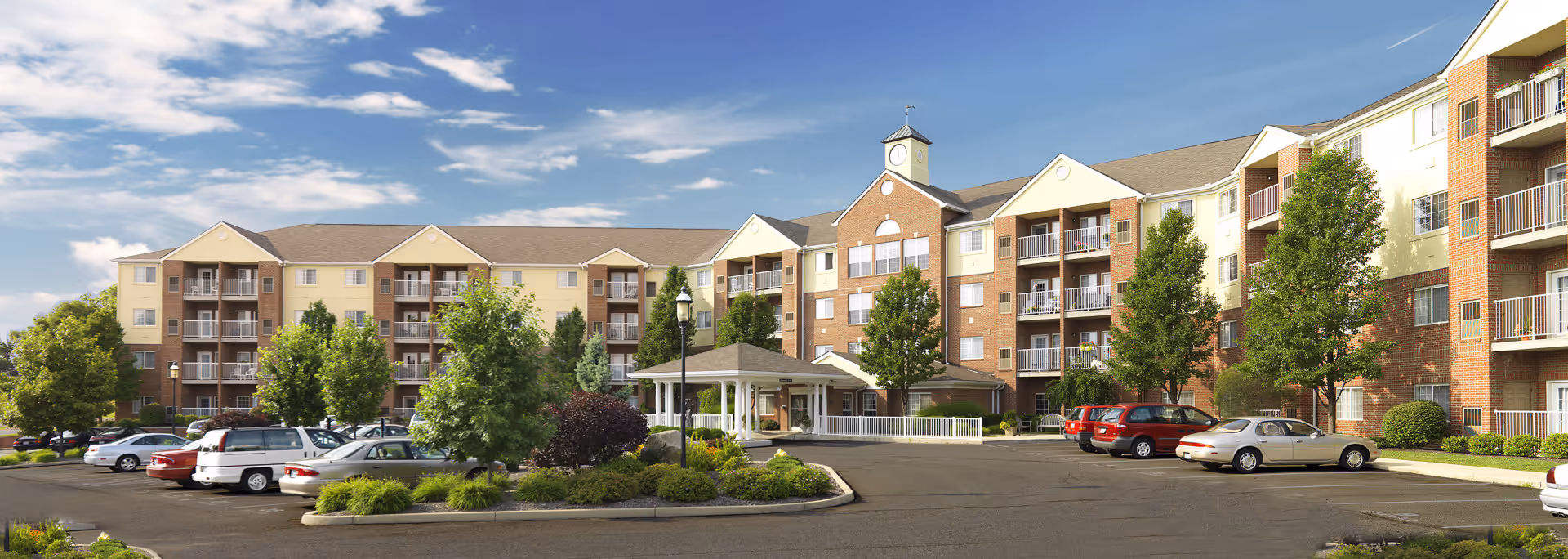 Exterior view of Barrington at Ft. Thomas, a multi-story senior living facility with brick and beige siding, balconies, a clock tower, surrounded by trees and a parking lot with several cars under a partly cloudy sky.
