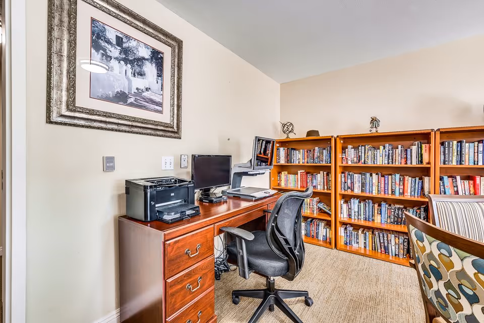 A cozy office or study area with a wooden desk holding a printer, computer monitor, and laptop. There is a black office chair in front of the desk. Behind the desk are three wooden bookshelves filled with books. A framed black and white picture hangs on the wall above the desk. The room has beige walls and carpeted flooring.