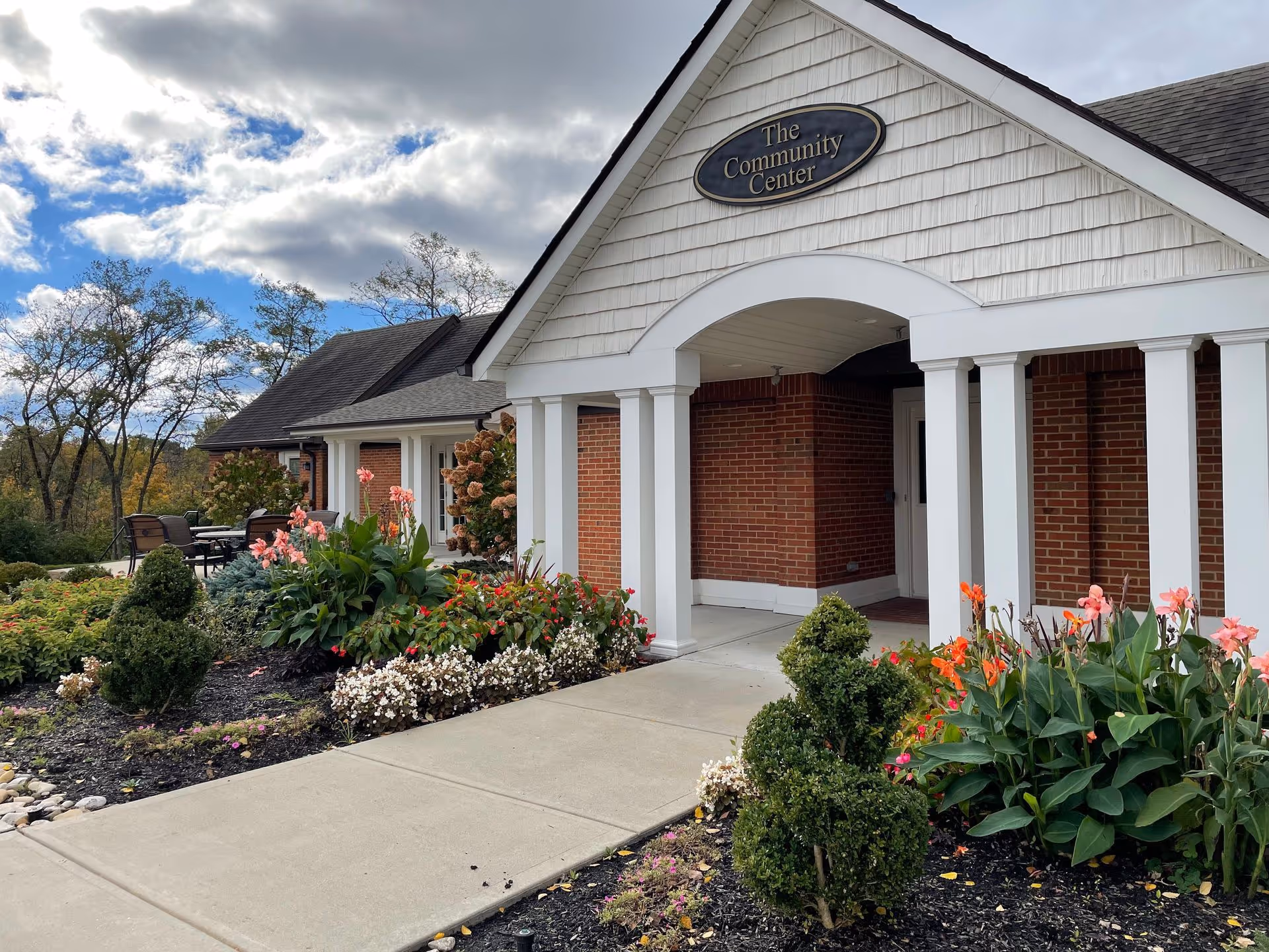 Exterior view of a community center building with white columns and brick walls, surrounded by landscaped flower beds with various colorful flowers and shrubs under a partly cloudy sky.