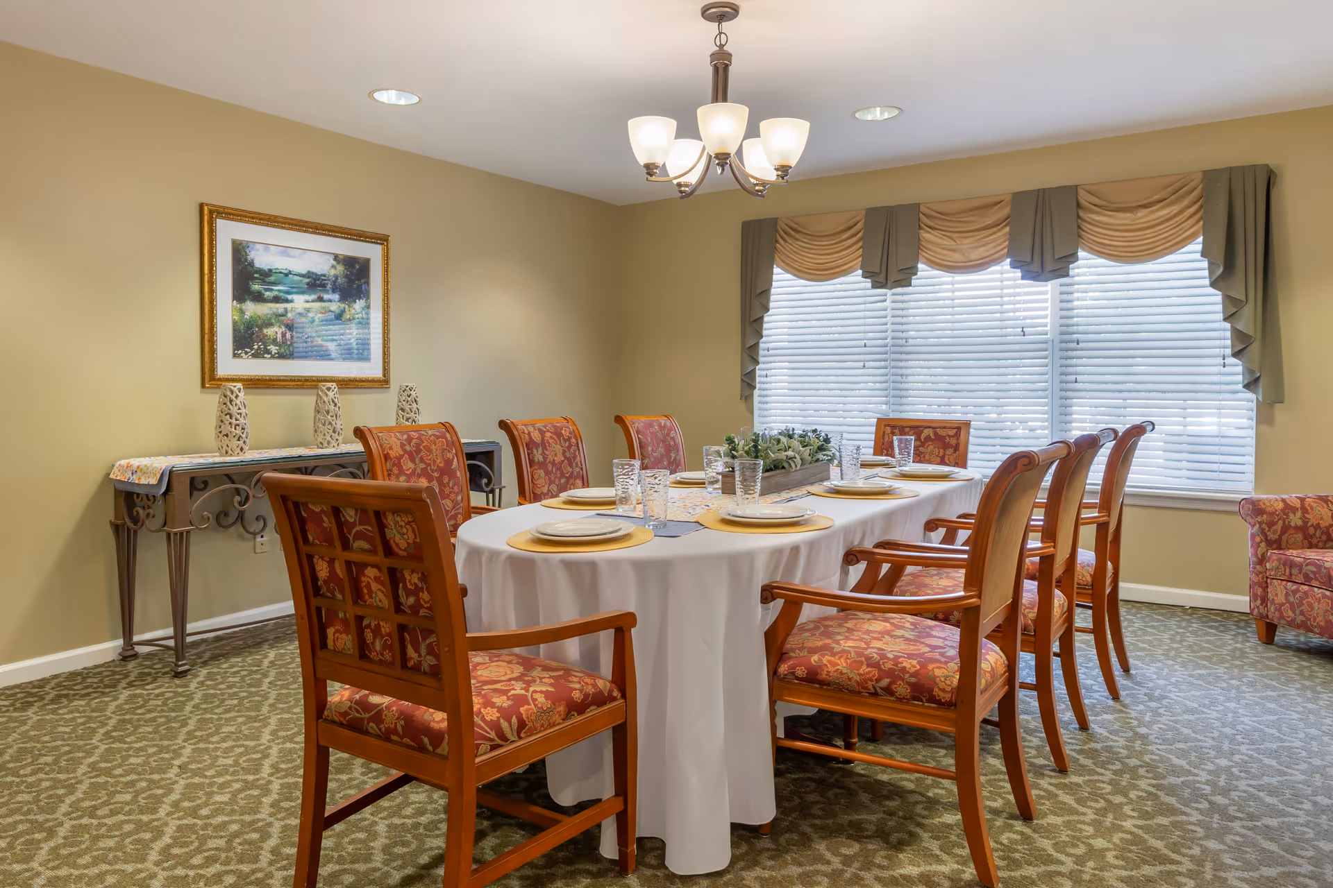 Dining room with a long table set for eight, patterned upholstered chairs, a console table with decor, wall art, and a large window with blinds and valances.