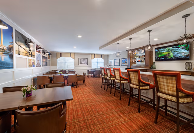 A spacious dining area with multiple tables and chairs, a bar counter with high chairs, and framed pictures on the walls. The room has a warm, inviting atmosphere with a red patterned carpet and soft lighting.