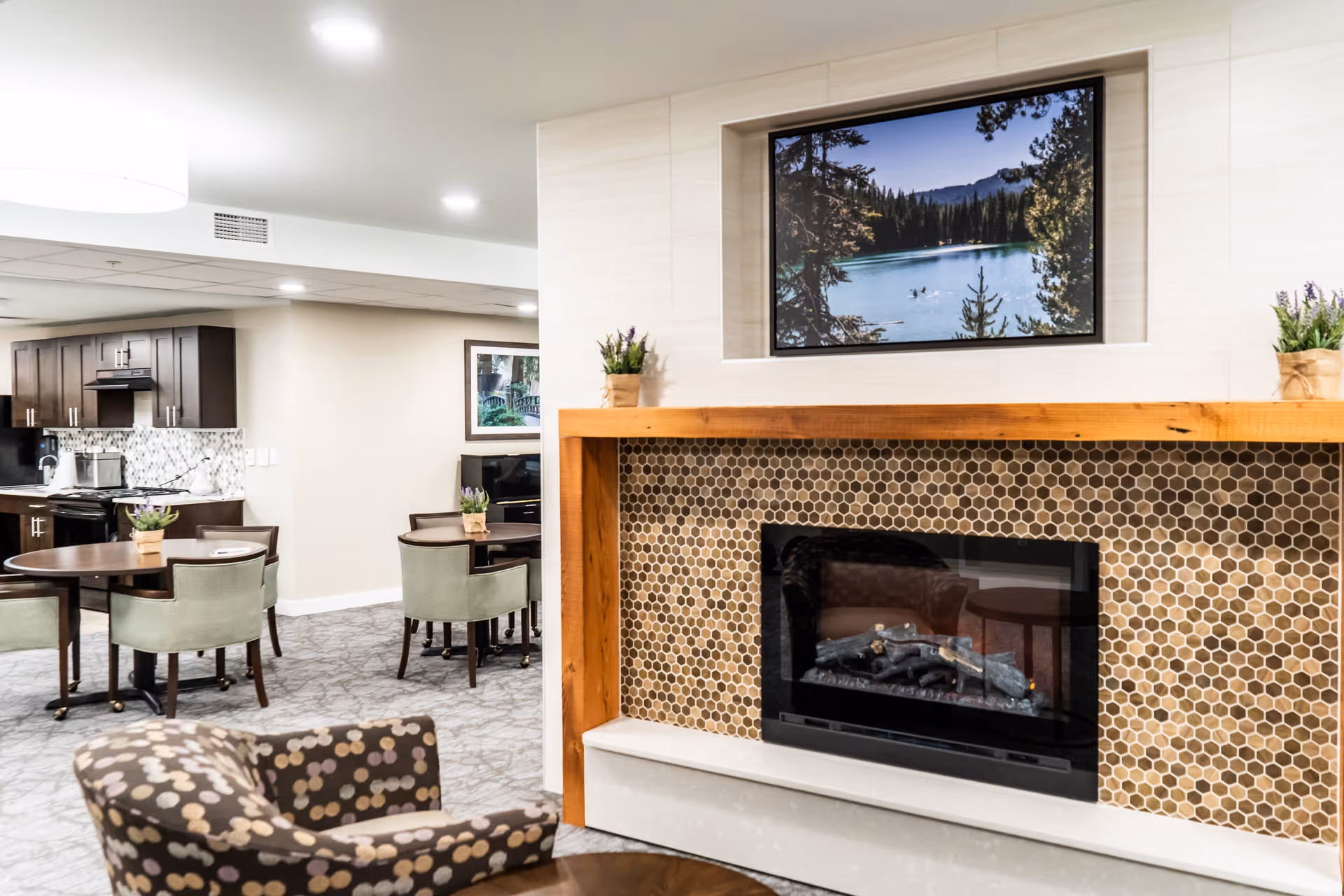 Communal living area with a tiled fireplace, framed art above, upholstered chairs, dining tables, and a kitchenette in the background.