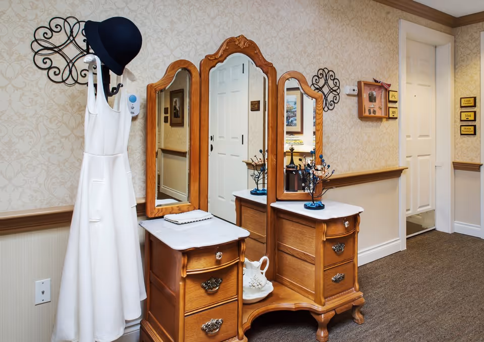 Wooden vanity with three mirrors and drawers in a decorated hallway next to a coat rack holding a white dress and black hat.