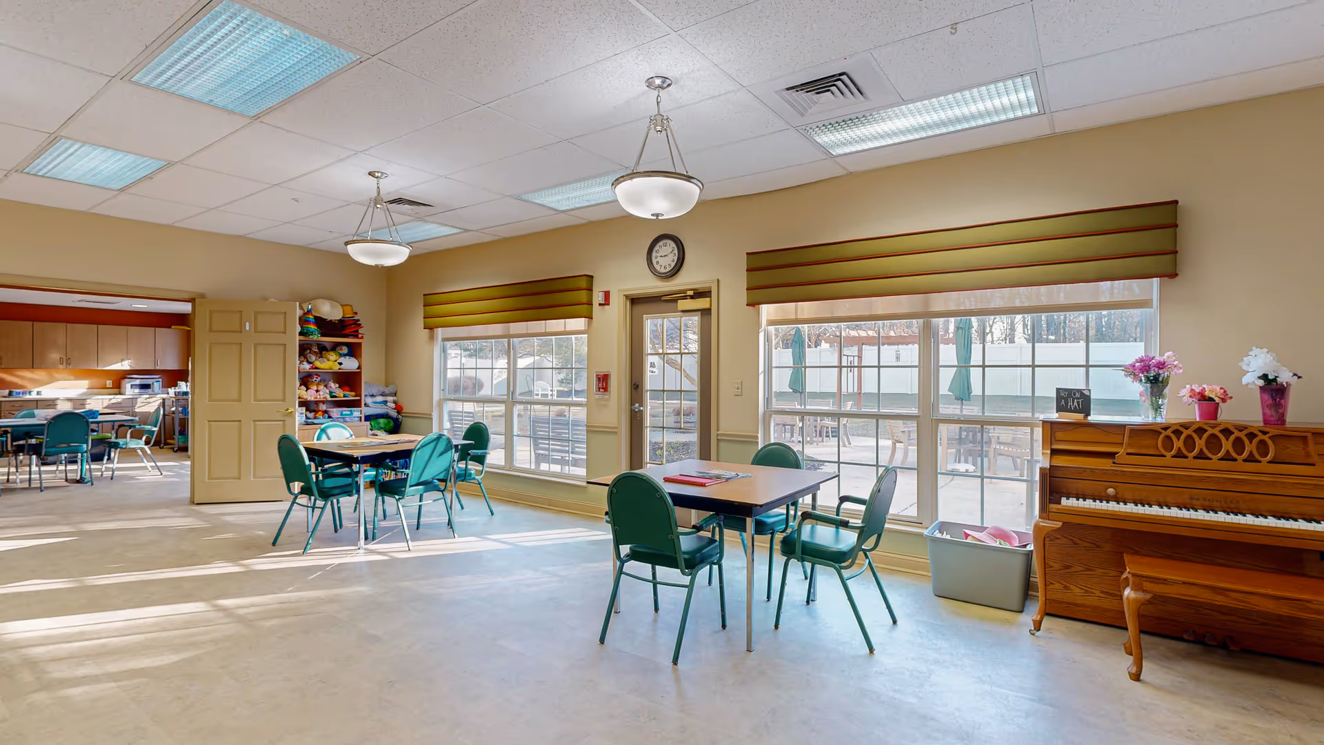 Well-lit communal activity/dining room with tables and green chairs, a piano, and large windows overlooking an outdoor patio.