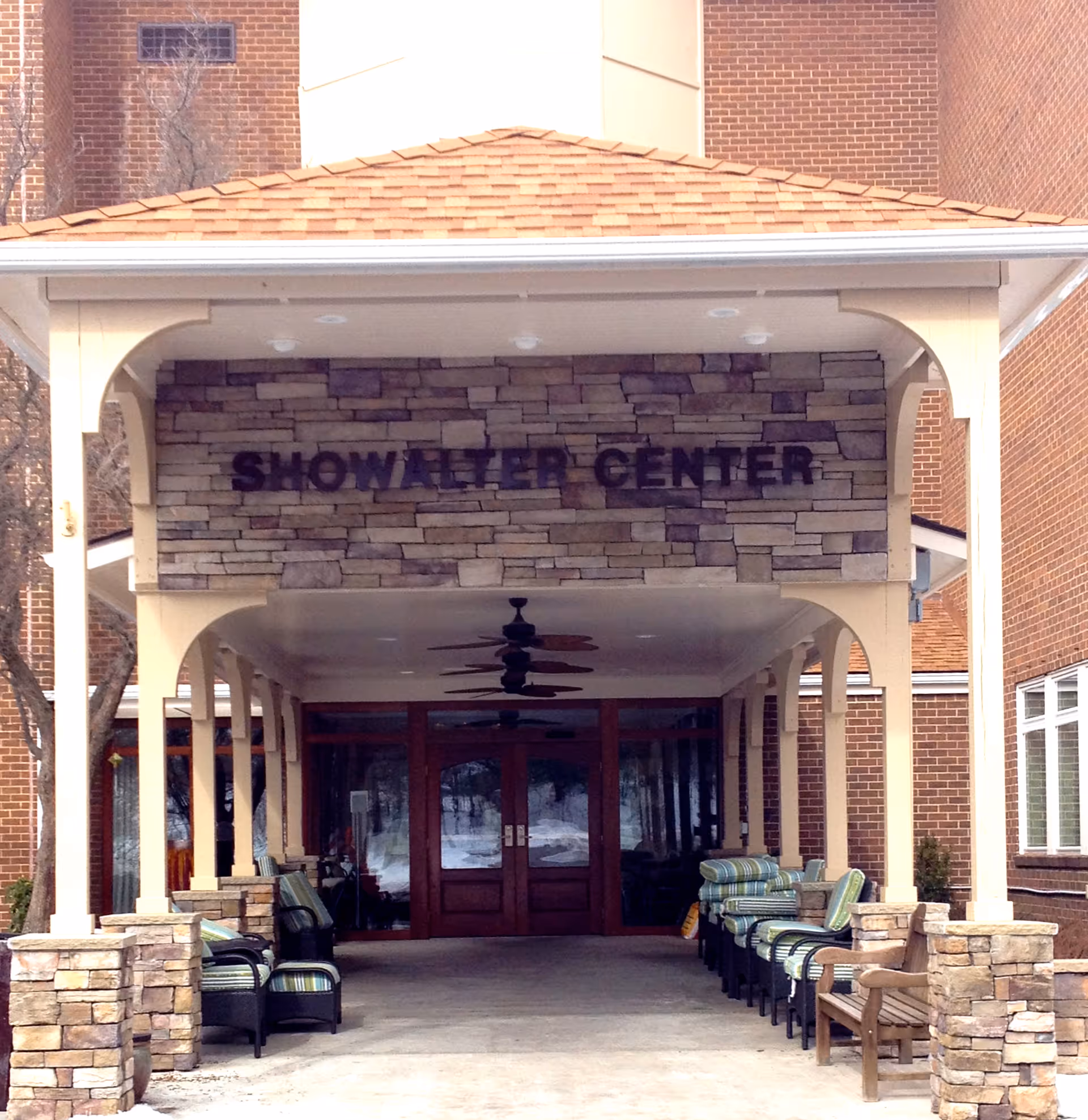 Covered entrance to the Showalter Center at Warm Hearth Village, featuring stone pillars and walls, a shingled roof, ceiling fans, and seating with cushioned chairs and benches along the sides.