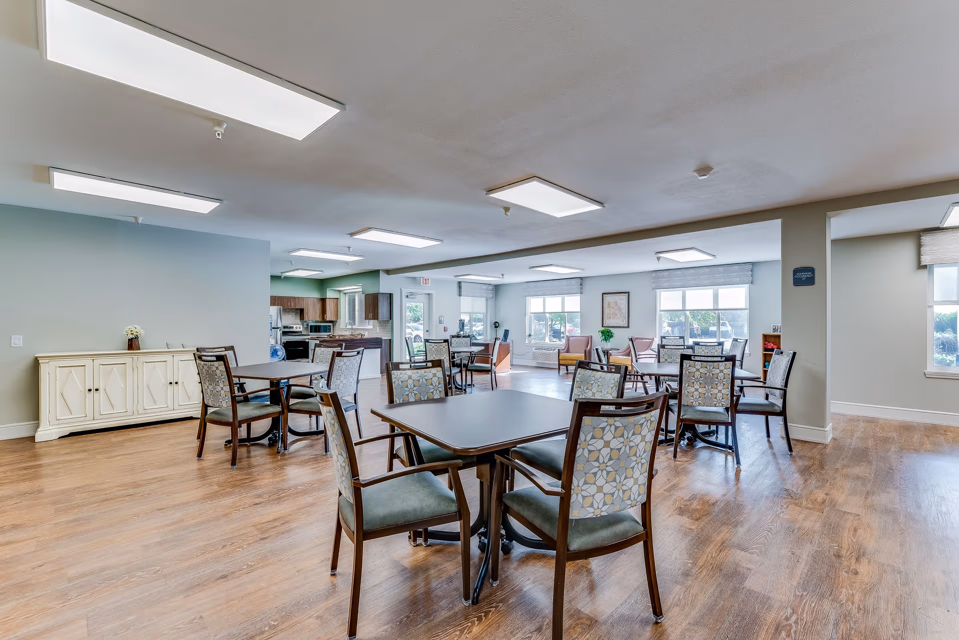 A bright and spacious dining area in a senior living facility with multiple tables and chairs arranged neatly on a wooden floor. Large windows allow natural light to fill the room, and there is a kitchen area visible in the background. The walls are painted in light colors, and the ceiling has several rectangular light fixtures.