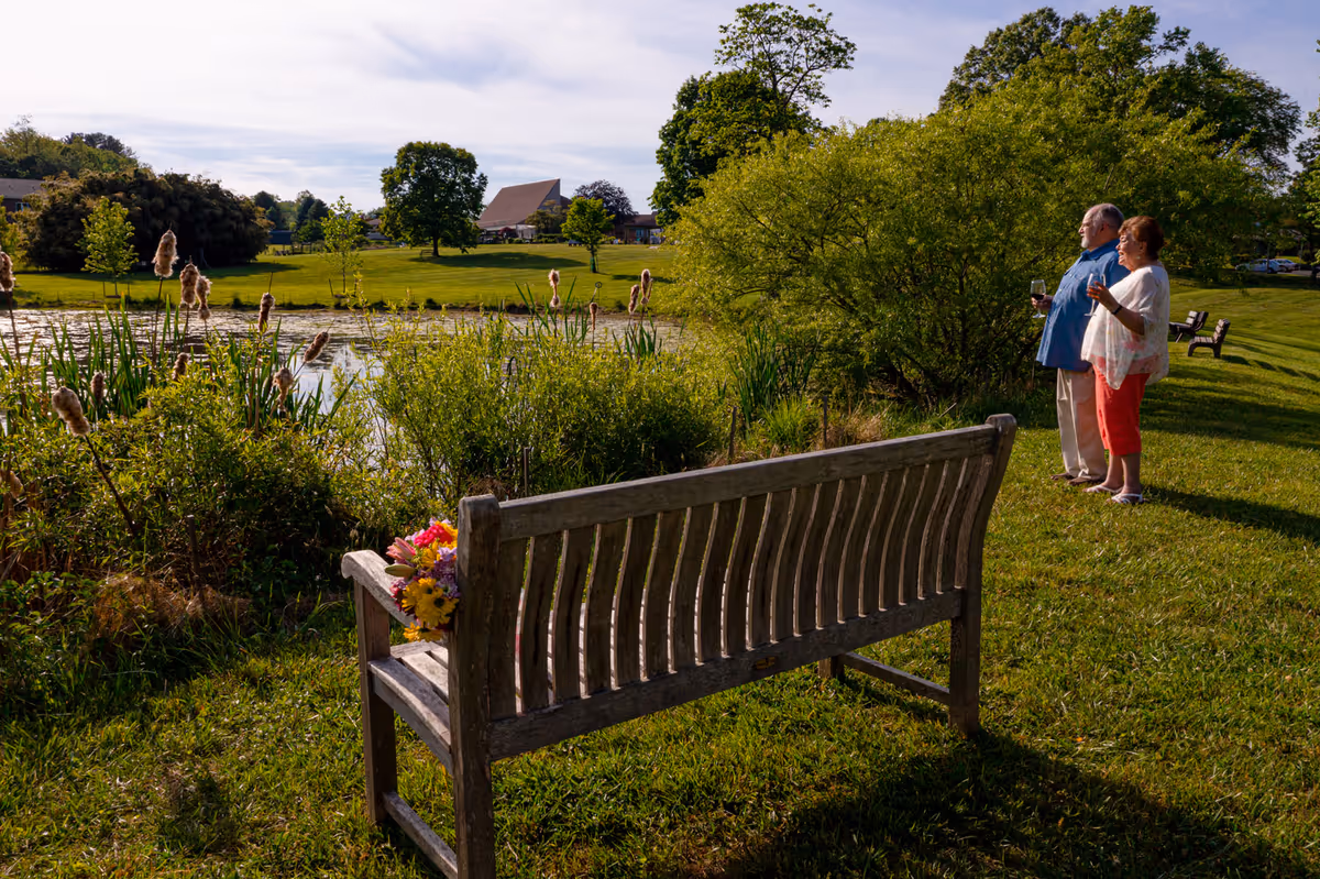 A wooden bench with flowers faces a pond while an older couple stands nearby holding drinks and enjoying the lakeside view.