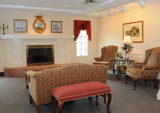 A cozy senior living common area with patterned upholstered chairs and a matching sofa, a red cushioned bench, a fireplace with a decorative mirror and framed pictures above it, a window with a red valance, and a side table with a lamp and plant.