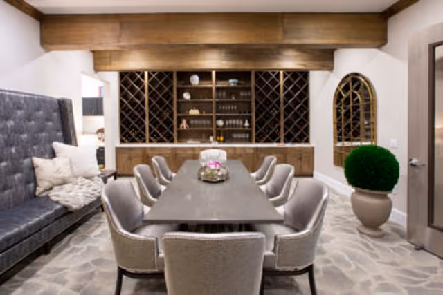 A dining room with a long rectangular table surrounded by eight upholstered chairs. On the left side, there is a tufted bench with pillows and a throw blanket. The back wall features wooden cabinetry with wine racks and shelves holding glassware and decorative items. A large mirror and a potted plant are on the right side. The room has a neutral color palette with wooden ceiling beams and a patterned carpet.