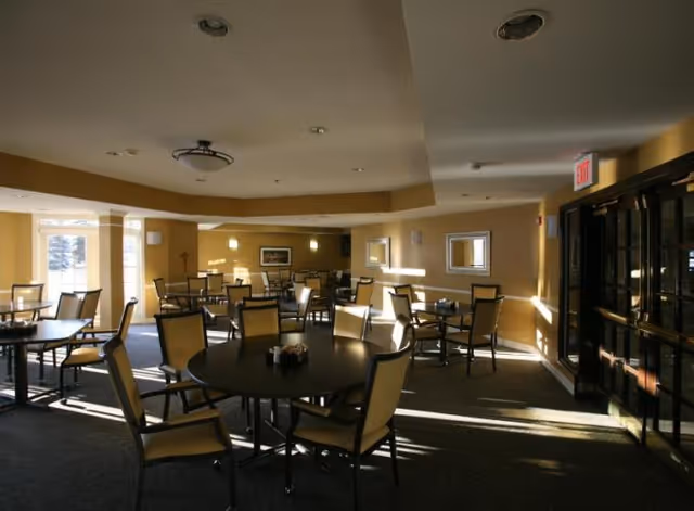 A dining room with multiple round tables and chairs arranged neatly. The room has beige walls, carpeted floors, and several windows letting in natural light. There are ceiling lights and wall-mounted lights, with an exit door visible on the right side.