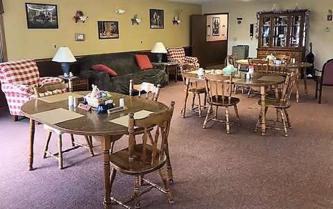 A cozy common area with several wooden tables and chairs arranged on a carpeted floor. There are two checkered armchairs, a dark green couch with red cushions, and a wooden cabinet with glass doors displaying items. The walls are decorated with framed pictures and sconces, and there are table lamps on side tables near the seating.