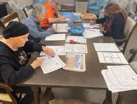 Four elderly individuals sitting around a table engaged in coloring and drawing activities with various coloring sheets, markers, and scissors spread out on the table in a communal room.