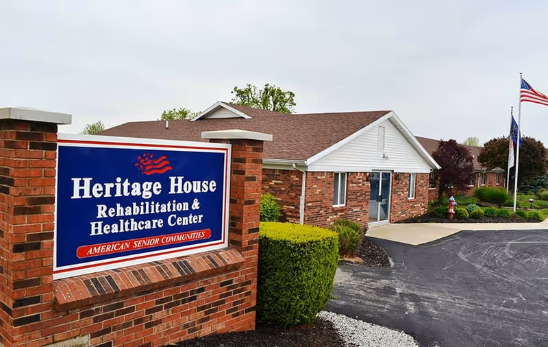 Exterior view of Heritage House Rehabilitation & Healthcare Center showing a brick sign with the facility name and a single-story brick building with a driveway, landscaping, and flags.