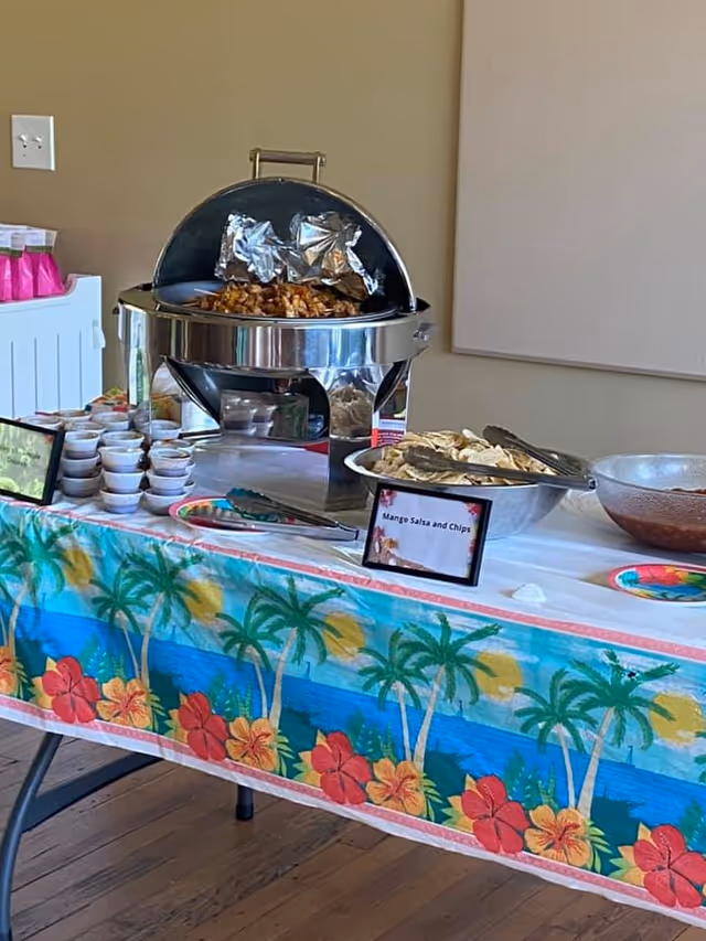 A buffet table with a tropical-themed tablecloth featuring palm trees and hibiscus flowers. On the table, there is a chafing dish with cooked food, a large bowl of chips with tongs, a bowl of salsa, and several small cups of sauce. Two small signs label the food items, one of which reads 'Mango Salsa and Chips'. The setting appears to be indoors with a beige wall and wooden floor.