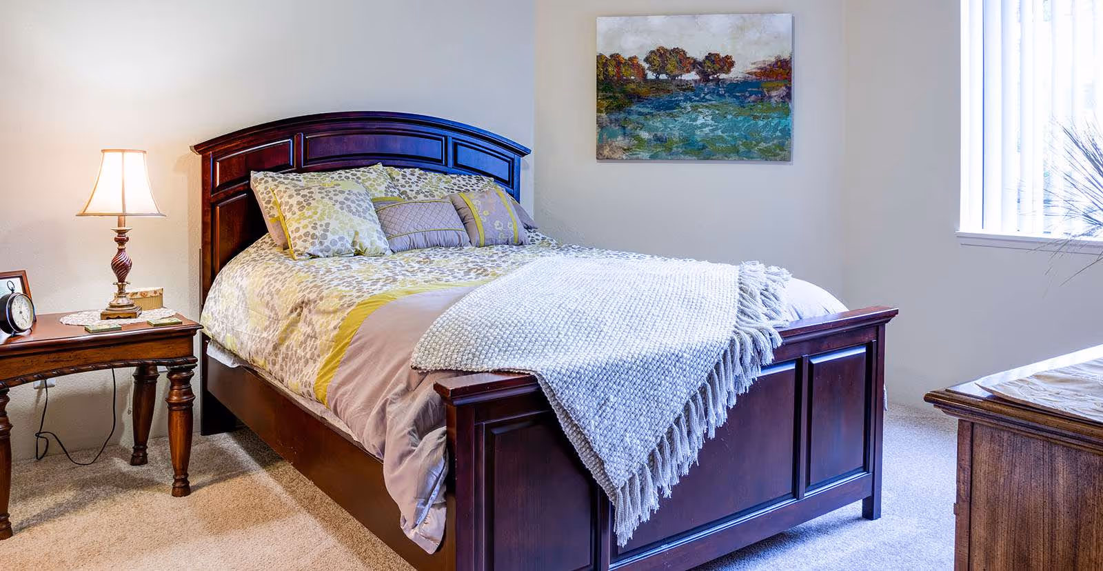 A cozy bedroom with a wooden bed frame and a neatly made bed featuring patterned pillows and a light-colored blanket. There is a wooden nightstand with a lamp, clock, and small decorative items. A colorful painting hangs on the wall above the bed, and a window with vertical blinds allows natural light into the room.