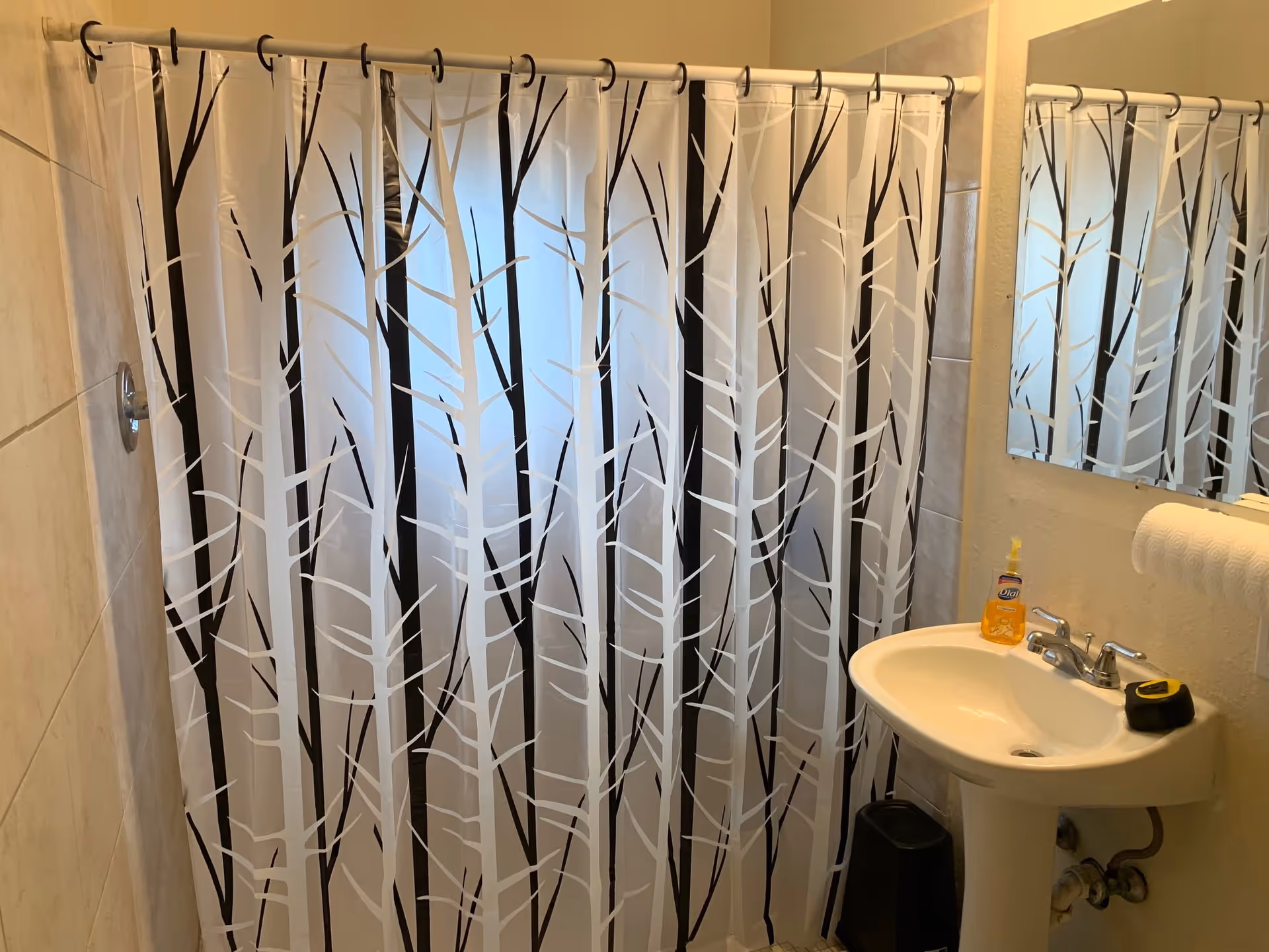 A bathroom with a white shower curtain featuring a black and white tree branch pattern. There is a white pedestal sink with a bottle of orange liquid soap and a black and yellow tape measure on it. A mirror is mounted on the wall above the sink, and a white towel is hanging on a towel rack next to the sink. The walls are tiled and painted beige.