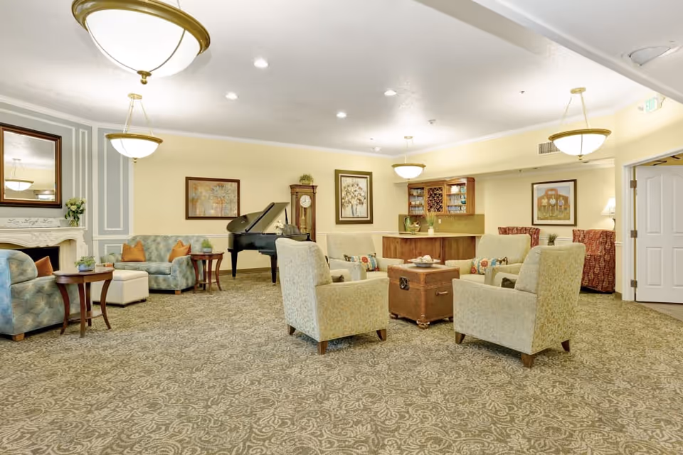 A spacious and well-lit living room area with patterned carpet, several upholstered armchairs and sofas arranged around a central wooden chest used as a coffee table. The room features a grand piano, a grandfather clock, framed artwork on the walls, and a small bar area with glassware. There are multiple ceiling lights and a large mirror above a fireplace on the left side.