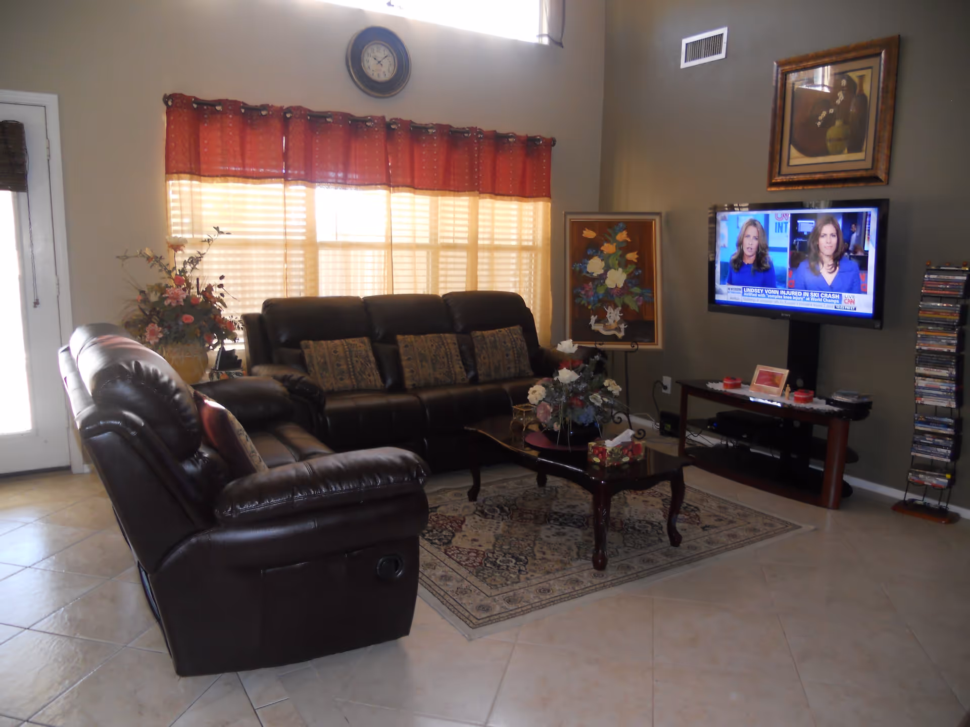A cozy living room with two dark brown leather sofas arranged around a wooden coffee table with floral decorations. A patterned rug lies underneath the table. A flat-screen TV is mounted on the wall showing a news broadcast. The room has beige tiled flooring, a window with red and beige curtains, a wall clock above the window, and framed artwork on the walls.