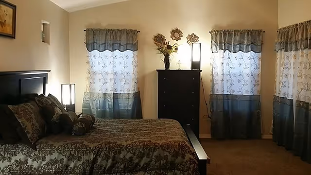 A bedroom with a large bed covered in patterned bedding and multiple pillows. There are three windows with sheer curtains allowing light to filter in. A tall black dresser with decorative flowers and a lamp on top is positioned between two of the windows. The walls are painted a light color and the floor is carpeted.