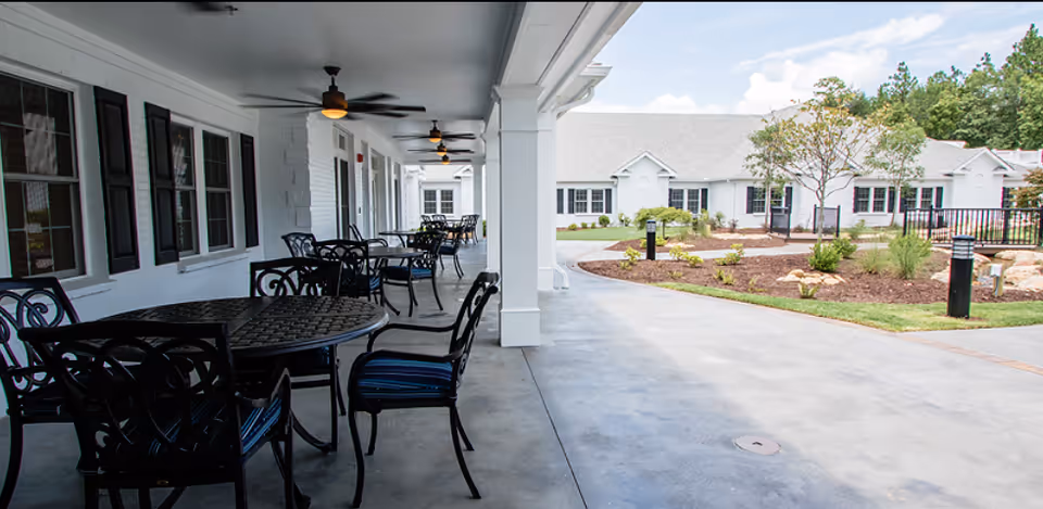 Covered outdoor patio with metal tables and chairs overlooking a landscaped courtyard and a single-story white building.
