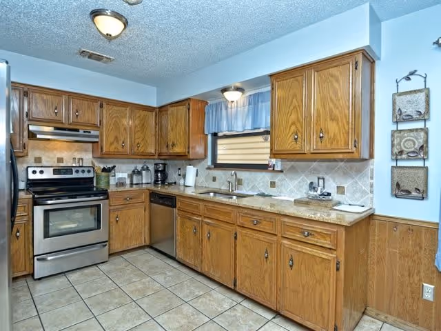 A kitchen with wooden cabinets, a stainless steel oven and dishwasher, a tiled floor, and a window with a blue valance above the sink. The countertops are light-colored, and there are various kitchen items such as a coffee maker and canisters on the counter.