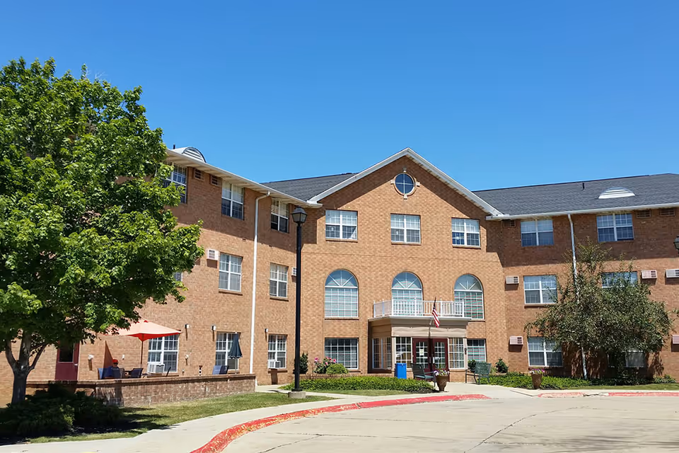 Exterior view of a three-story brick building with multiple windows, a small balcony, and an American flag near the entrance. There are trees and outdoor seating with umbrellas on the left side, and a clear blue sky above.