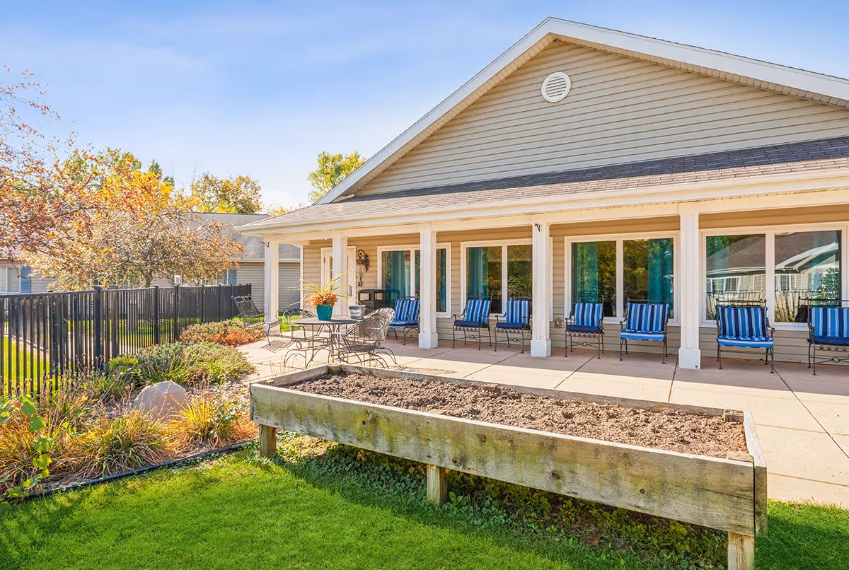 Outdoor patio area of a senior living facility with a raised garden bed in the foreground, a table with chairs, and a row of blue-striped cushioned chairs lined up against the building. The building has beige siding and large windows, with a clear blue sky above and some trees with autumn foliage nearby.