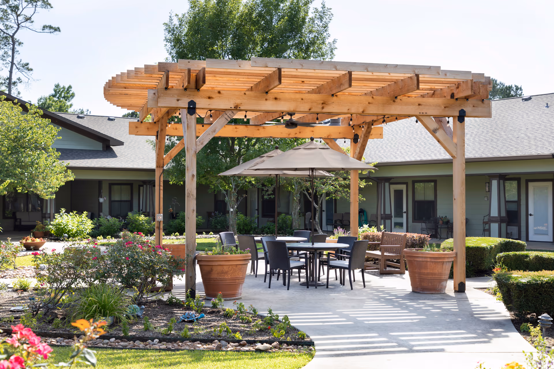 Outdoor seating area at Hawkins Creek Assisted Living and Memory Care featuring a wooden pergola with string lights, a round table with chairs, an umbrella, large potted plants, and surrounding garden beds with flowers and shrubs. The background shows a single-story building with multiple doors and windows.