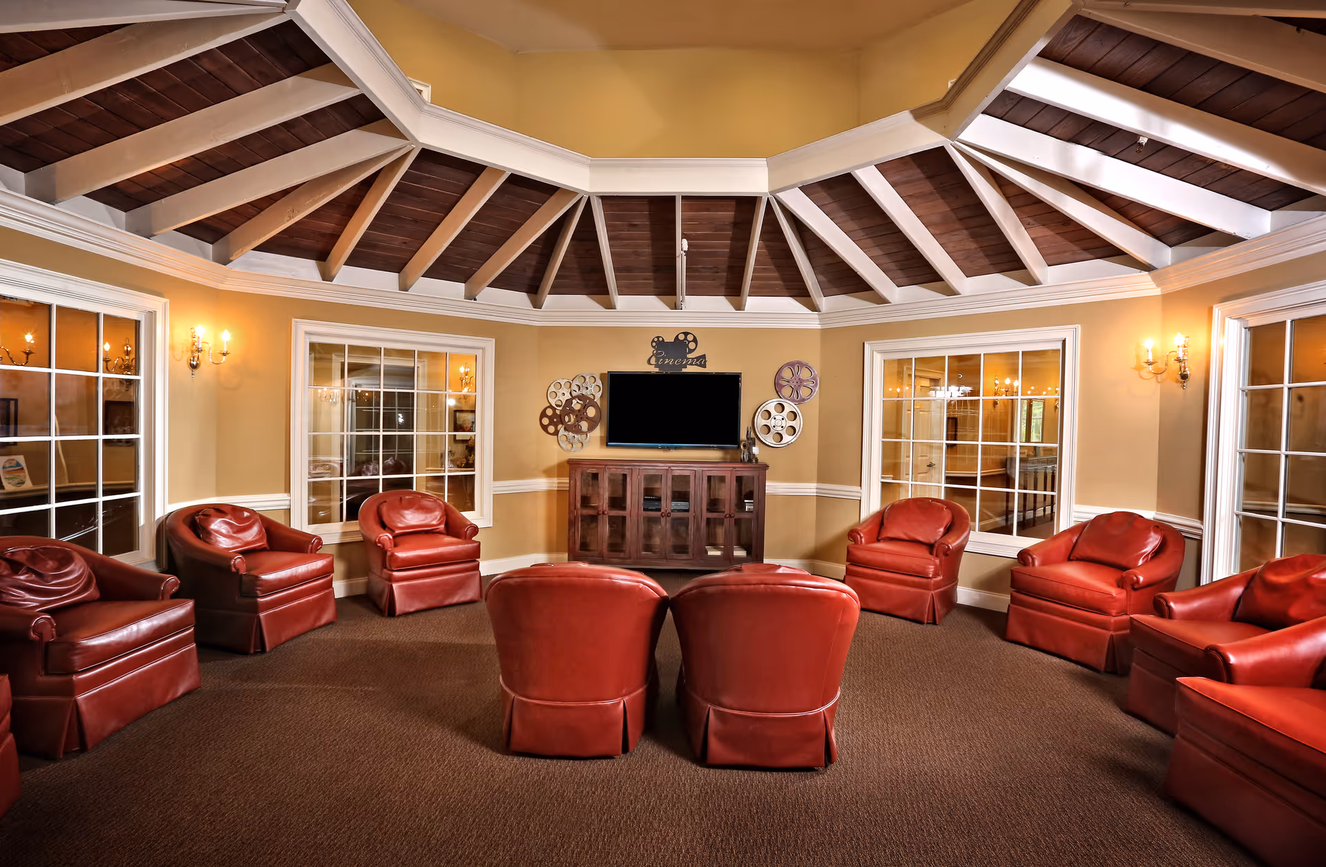A cozy senior living common area with a circular arrangement of red leather armchairs facing a flat-screen TV mounted on a wooden cabinet. The room features a high, wooden beamed ceiling with white trim, beige walls, and large windows with white frames. Decorative film reels and a 'Cinema' sign adorn the wall above the TV.