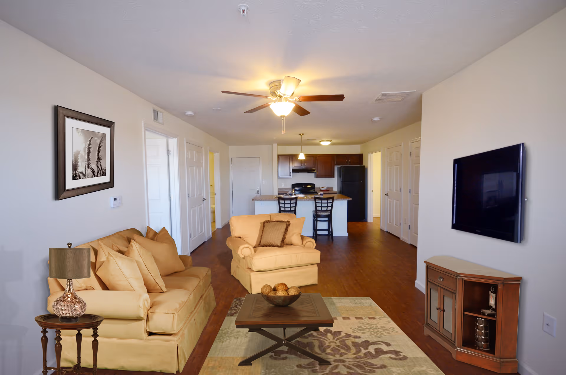 Interior view of a senior living apartment at The Reserve at Lakeview Landing featuring a living room with a beige sofa, armchair, wooden coffee table with decorative balls, side table with a lamp, wall-mounted flat screen TV, and a kitchen area with dark wood cabinets, black refrigerator, and two bar stools at the counter.