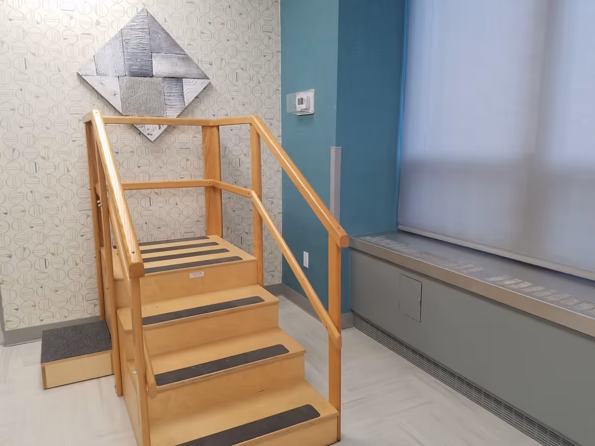 A small wooden staircase with handrails on both sides, designed for rehabilitation or physical therapy, placed indoors against a wall with patterned wallpaper and a blue accent wall. There is a metallic abstract wall decoration above the stairs and a large window with a white blind on the right side.