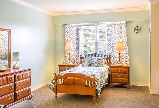 A cozy bedroom with a single wooden bed featuring patterned bedding and pillows. The room has two wooden nightstands with lamps on either side of the bed, a large window with patterned curtains letting in natural light, and a wooden dresser with a mirror on the left side. The walls are painted light green and the floor is carpeted.