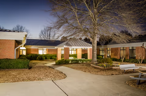 Exterior view of a brick senior living facility with a curved walkway, benches, and a large tree.
