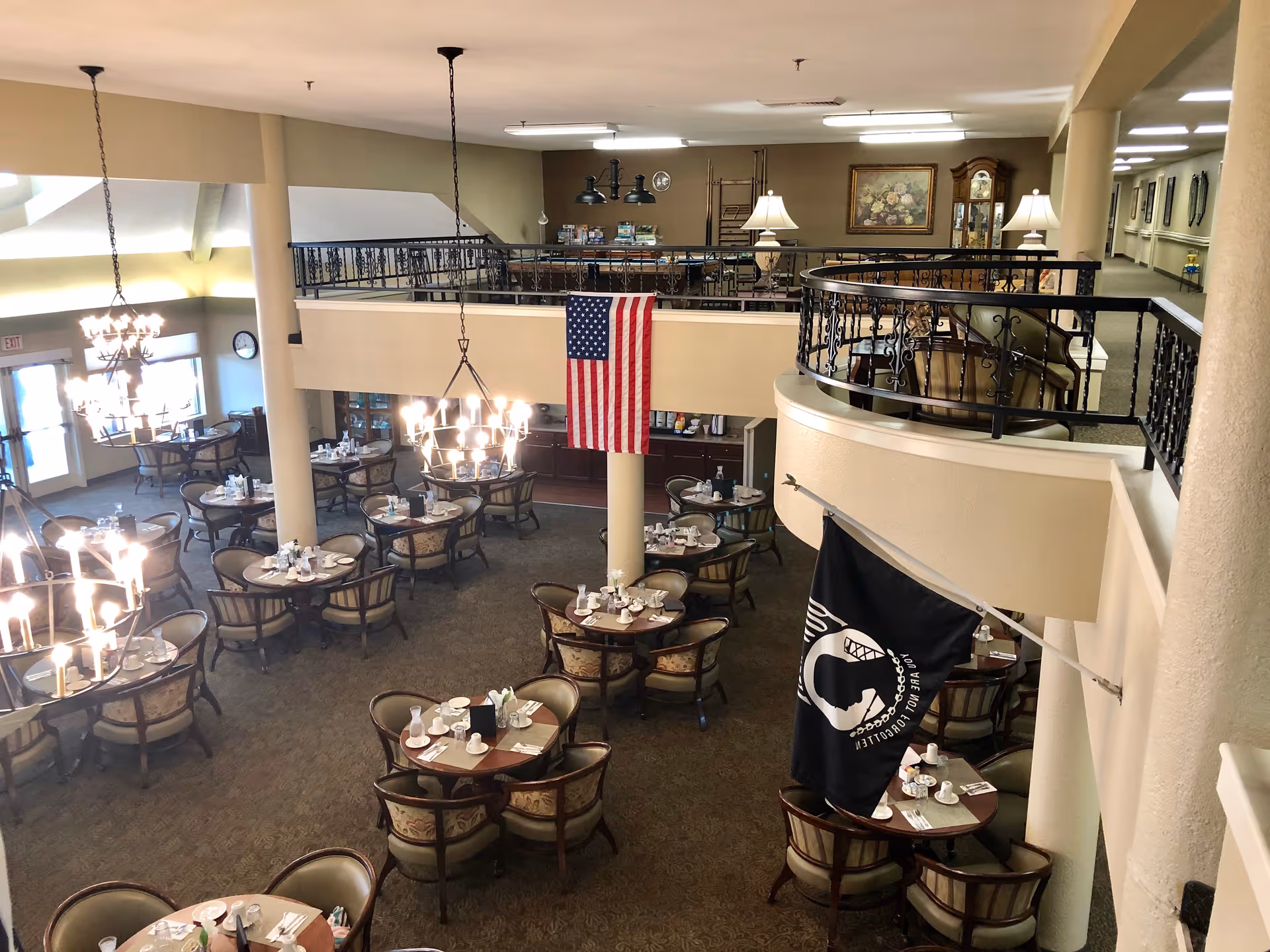 Interior view of a senior living facility dining room with multiple round tables set with cups, plates, and utensils. The room features chandeliers, an American flag hanging from the upper level, and a black POW/MIA flag. The upper level has a railing with chairs and lamps, and there are paintings and a grandfather clock on the walls.