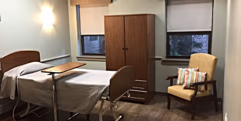 Hospital patient room with an adjustable bed, overbed table, wardrobe, and chair.