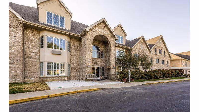 Exterior view of a large brick assisted living facility building with multiple windows, a covered entrance with an archway, benches near the entrance, and a paved driveway in front.