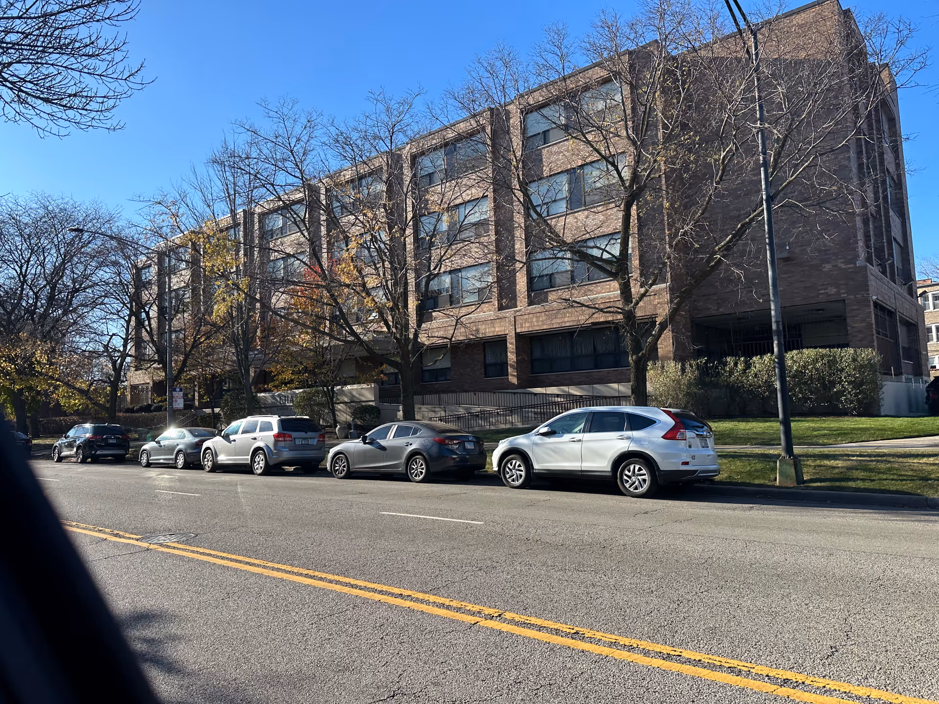 Exterior view of a multi-story brick building with several windows, situated along a street with parked cars and leafless trees under a clear blue sky.