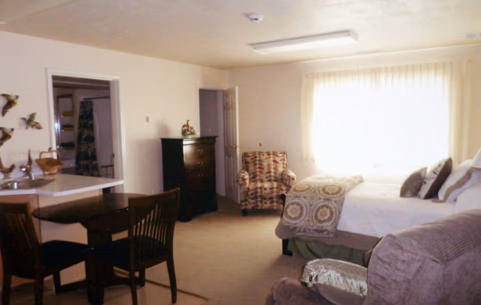 A cozy bedroom with a bed covered in patterned blankets and pillows, a cushioned armchair with a colorful leaf pattern, a dark wooden dresser, and a small dining table with two chairs. The room has beige walls and carpet, with a window covered by sheer curtains letting in natural light.