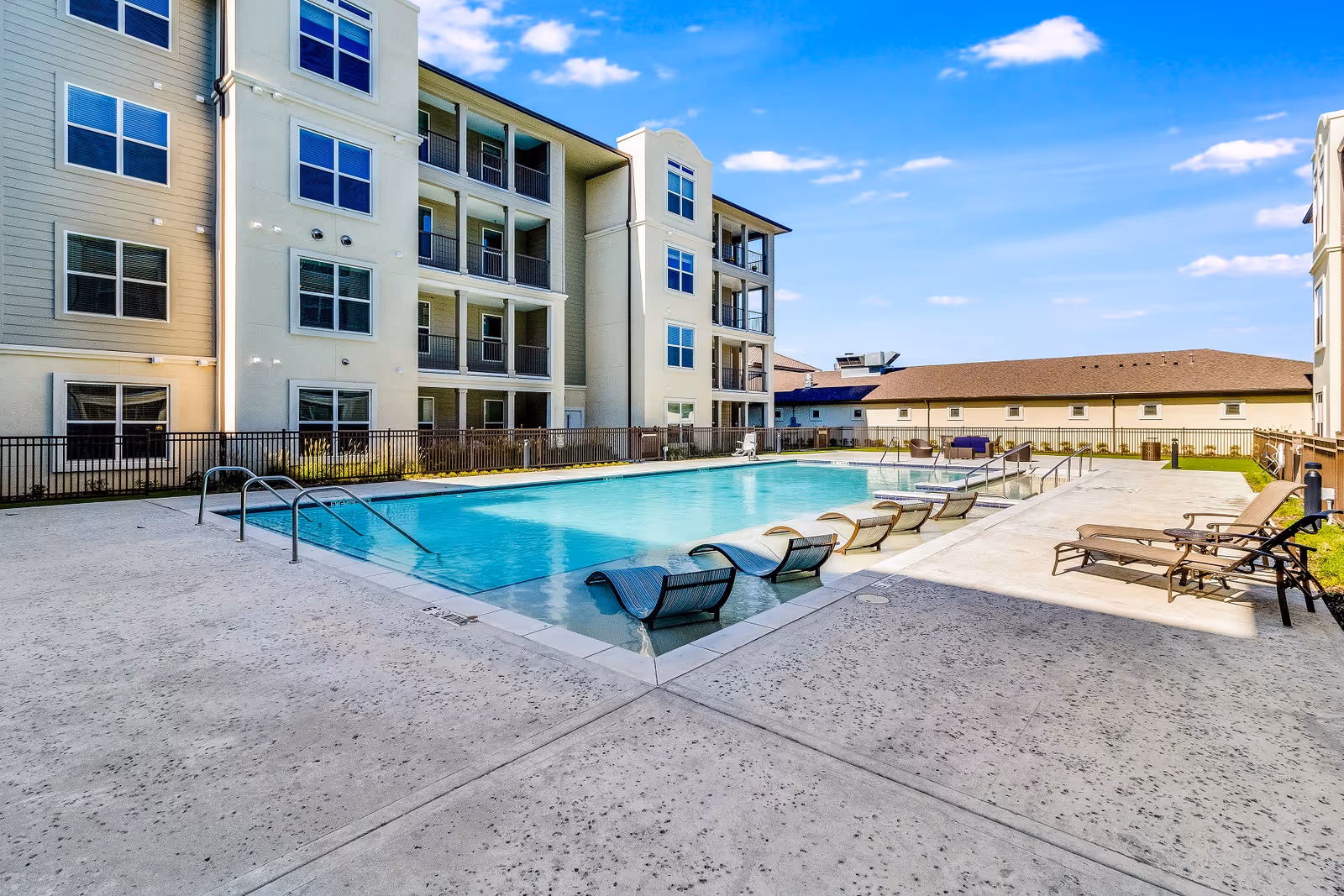 Outdoor swimming pool with lounge chairs beside a multi-story senior living building under a blue sky.