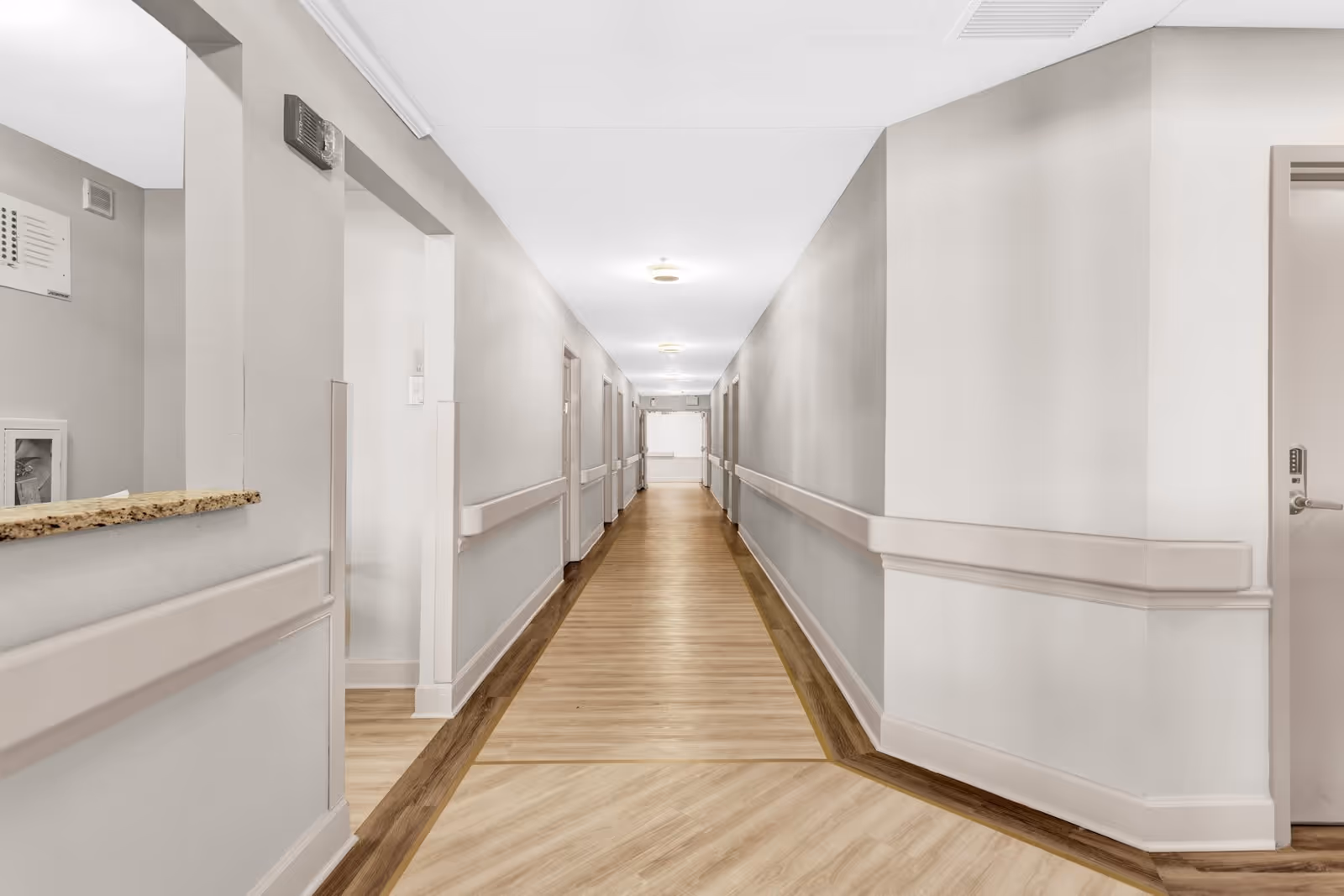 Well-lit long interior hallway in a senior living facility with handrails and wood-tone flooring.