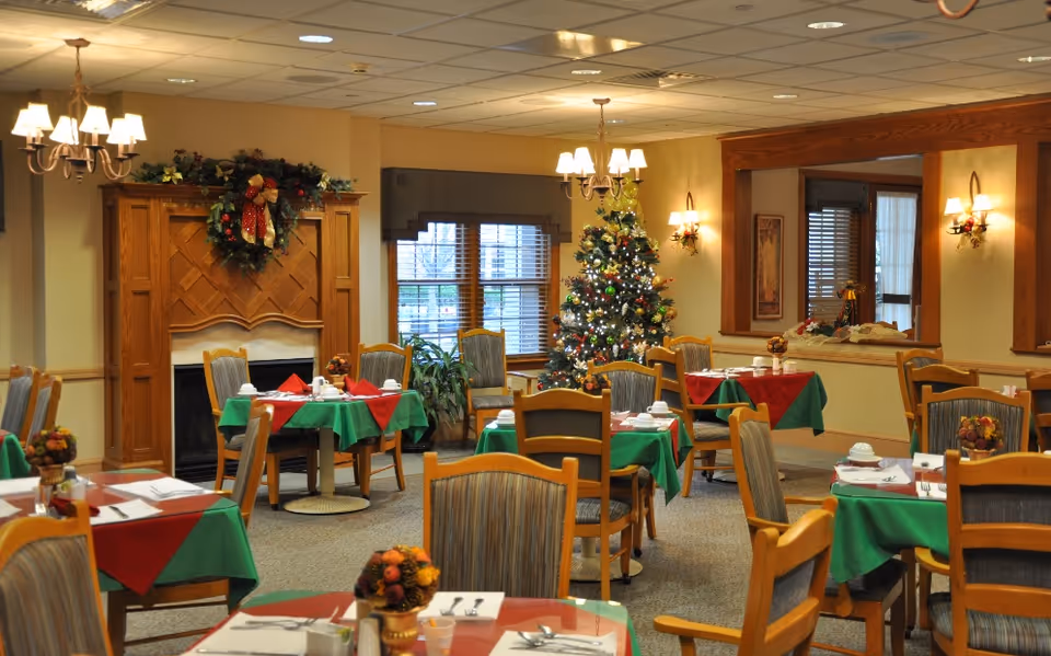 Dining room with tables set with red and green tablecloths, wooden chairs, a decorated Christmas tree, and a fireplace.