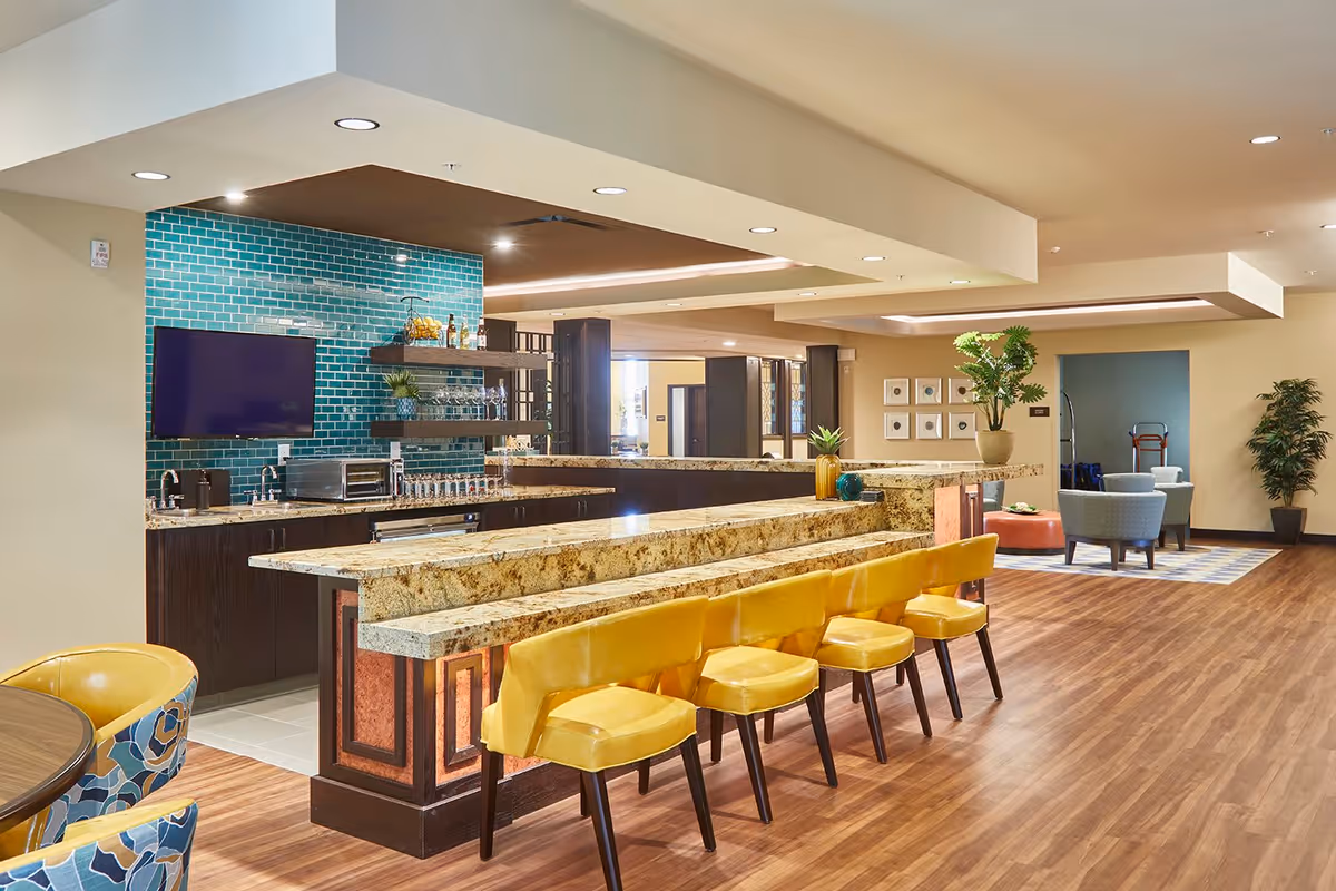 Interior view of a modern common area with a long granite countertop bar featuring six yellow chairs. Behind the bar is a kitchen area with dark cabinets, a teal tiled backsplash, a mounted flat-screen TV, and shelves with glassware and decorative items. The room has wood flooring, recessed lighting, and additional seating with blue and orange chairs in the background.