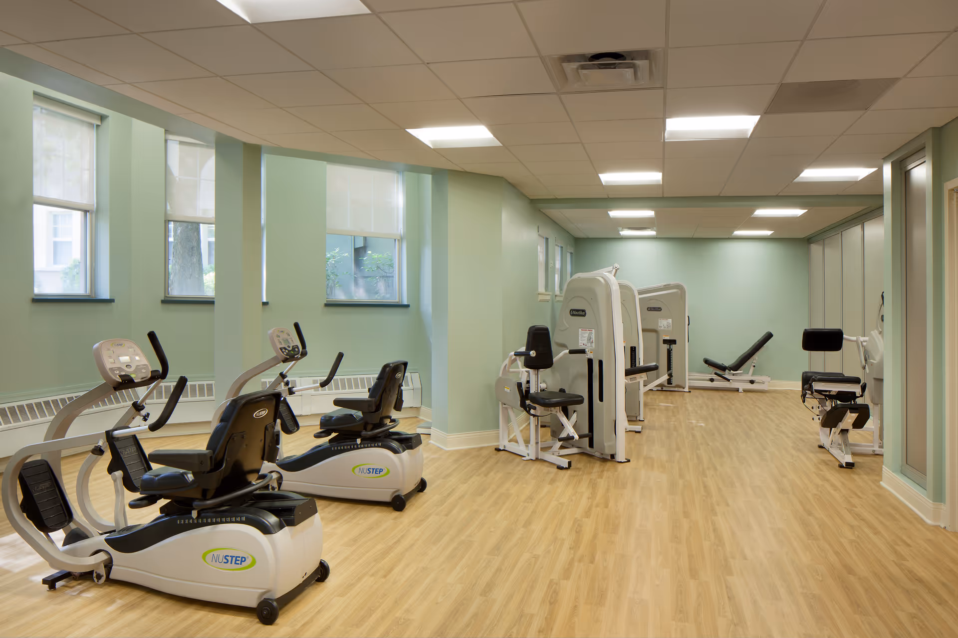 A fitness room with exercise equipment including two recumbent stationary bikes and several strength training machines. The room has light green walls, wood-patterned flooring, and several windows letting in natural light.