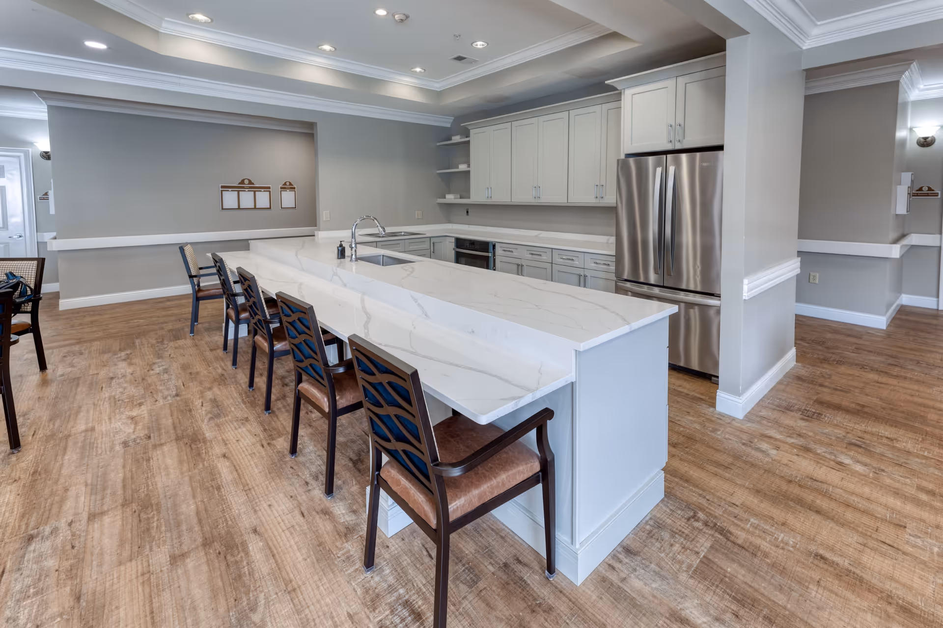 A modern kitchen area with a long white marble countertop island surrounded by six wooden chairs with cushioned seats. The kitchen features white cabinetry, stainless steel refrigerator, and built-in appliances. The floor is wooden, and the walls are painted light gray with white trim. The ceiling has recessed lighting.