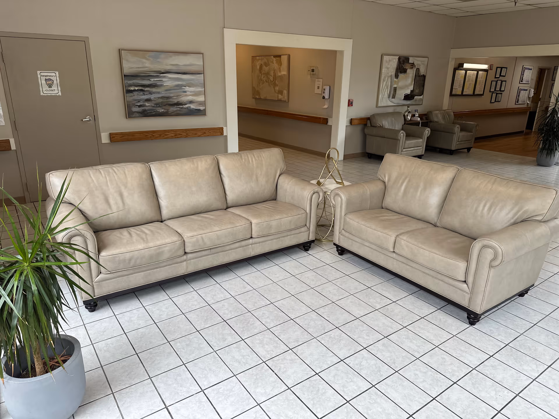 Lobby seating area with two beige sofas on a tiled floor, a potted plant, and additional chairs and artwork in the background.